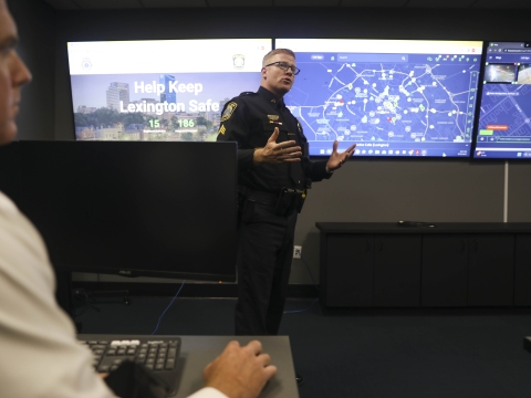 A police officer stands in a dark room with large computer monitors on the wall
