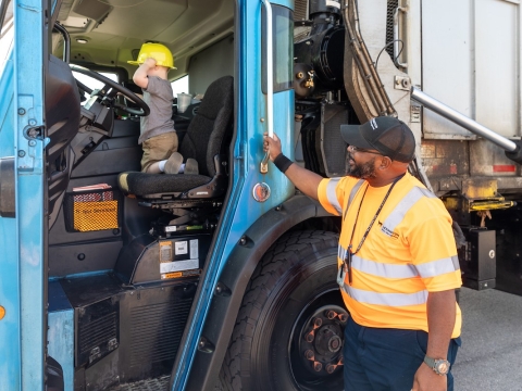 a driver standing next to a garbage truck, with a happy look on his face as he watches a small child sitting in the driver's seat of the truck
