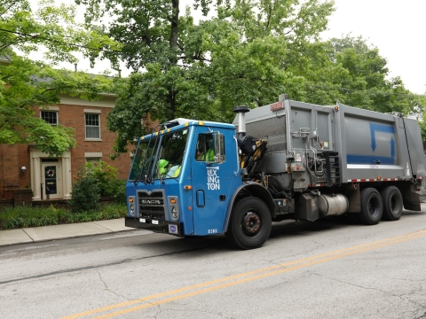 a trash truck driving down the road in front of a brick multifamily building with trees lining the sidewalk