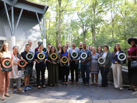 a group of about 2 dozen people standing outside with trees in the background, holding Green Check plaques