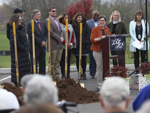 Mayor Linda Gorton speaks at a lectern with councilmembers behind her and a pile of dirt with shovels to their side